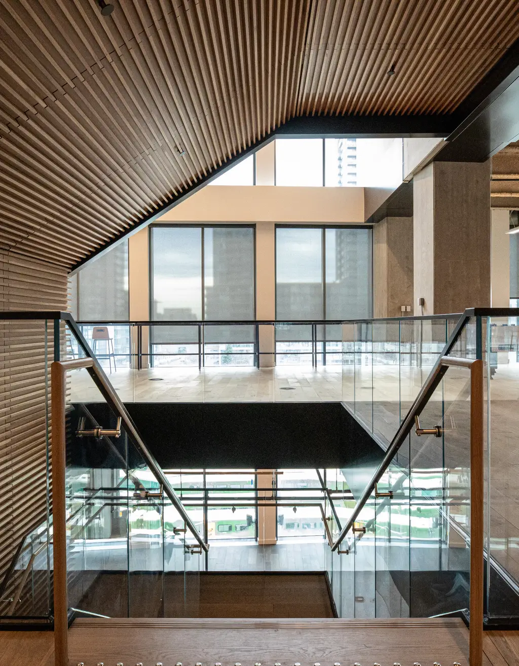 Office stairwell framed by a wood-slat ceiling and glass railings, with a city view through tall windows